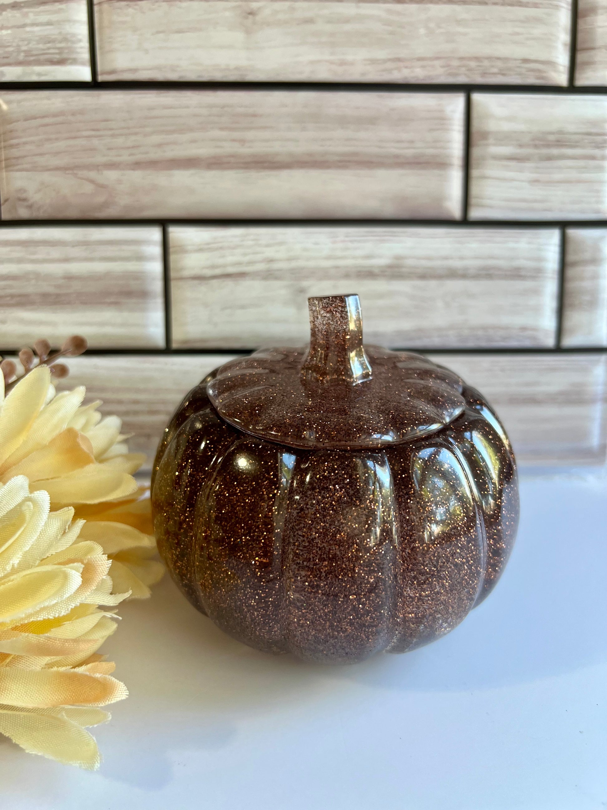 A brown glittery pumpkin-shaped container sits on a white surface, with a yellow flower to the left.