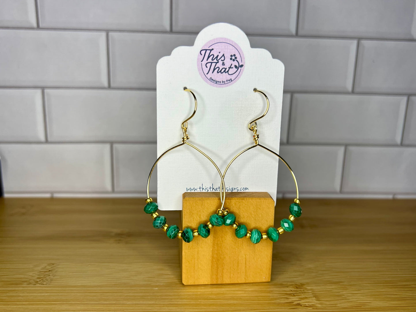 A pair of gold hoop earrings with green gemstone beads, displayed on a wooden stand against a white tiled wall.
