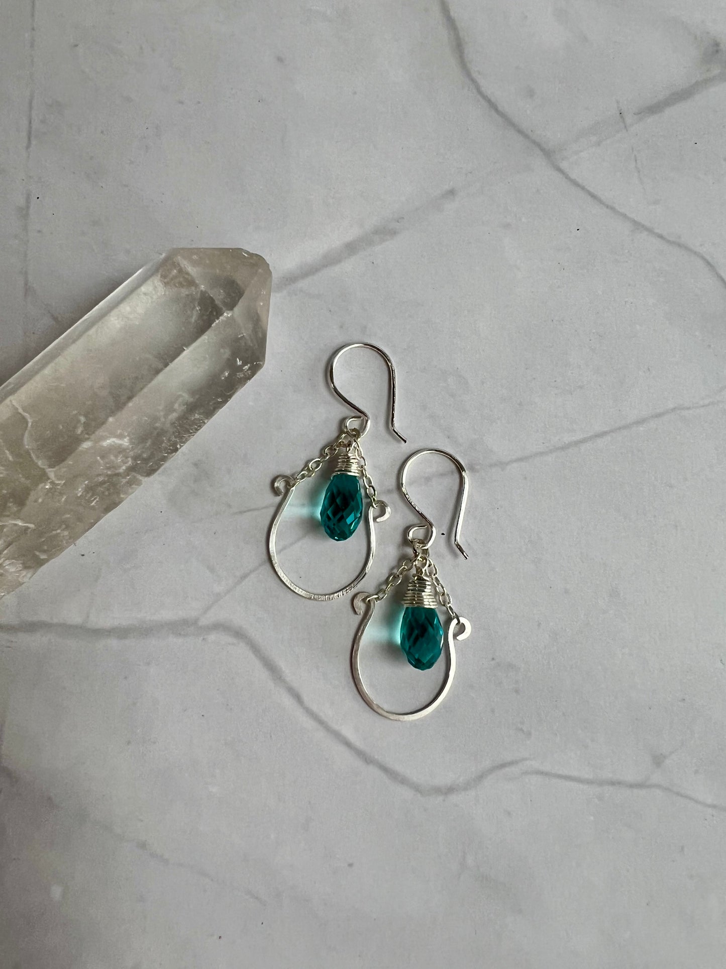 A pair of silver earrings with turquoise stones, resting on a marble surface next to a crystal.
