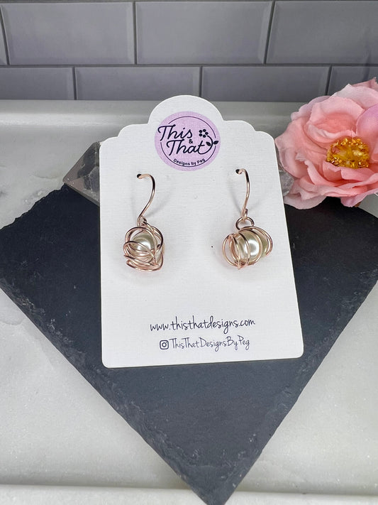 This image shows a pair of earrings displayed on a white card, with a pink flower in the background.