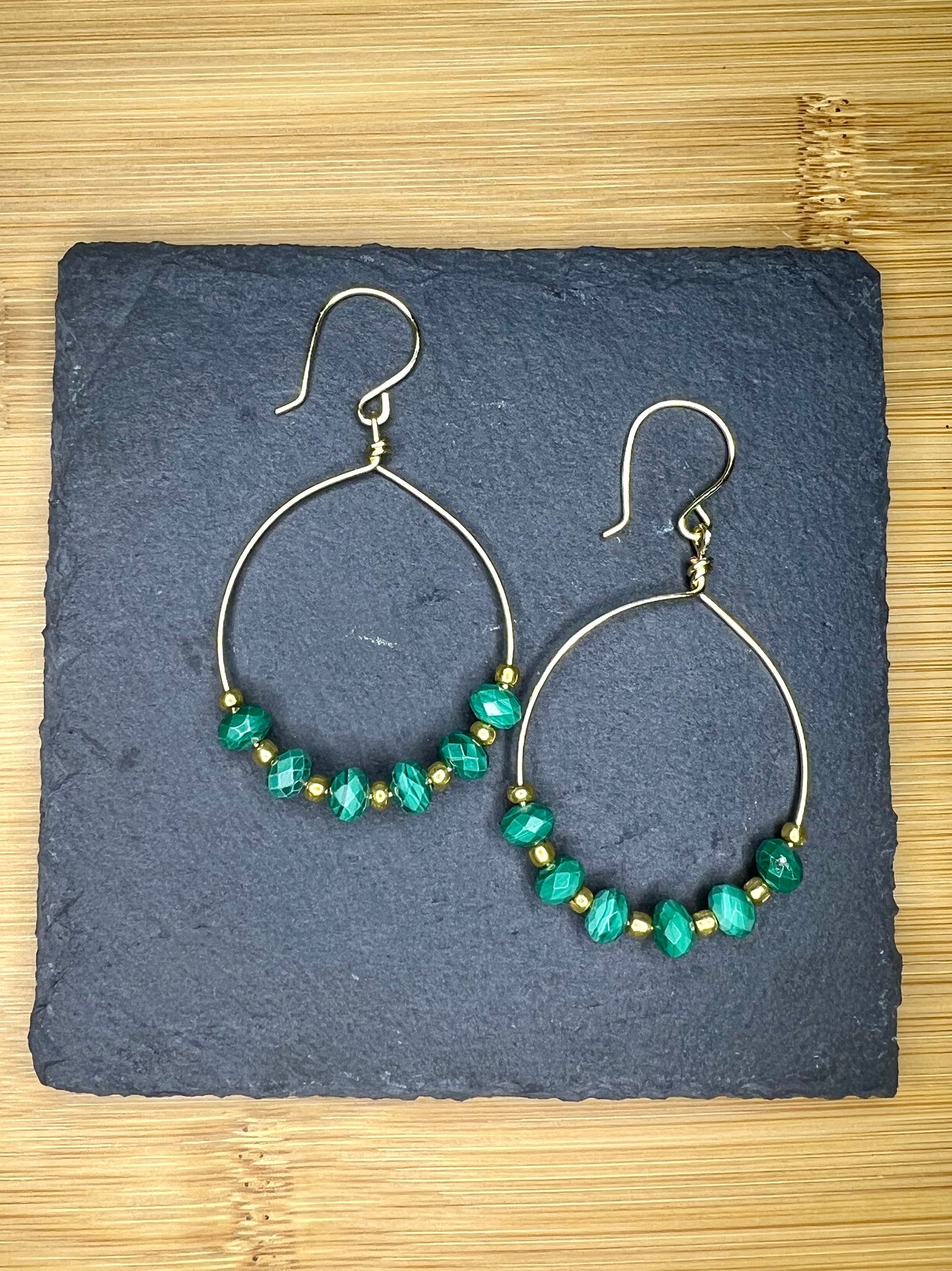 A pair of gold hoop earrings with green beads, displayed on a dark slate surface.