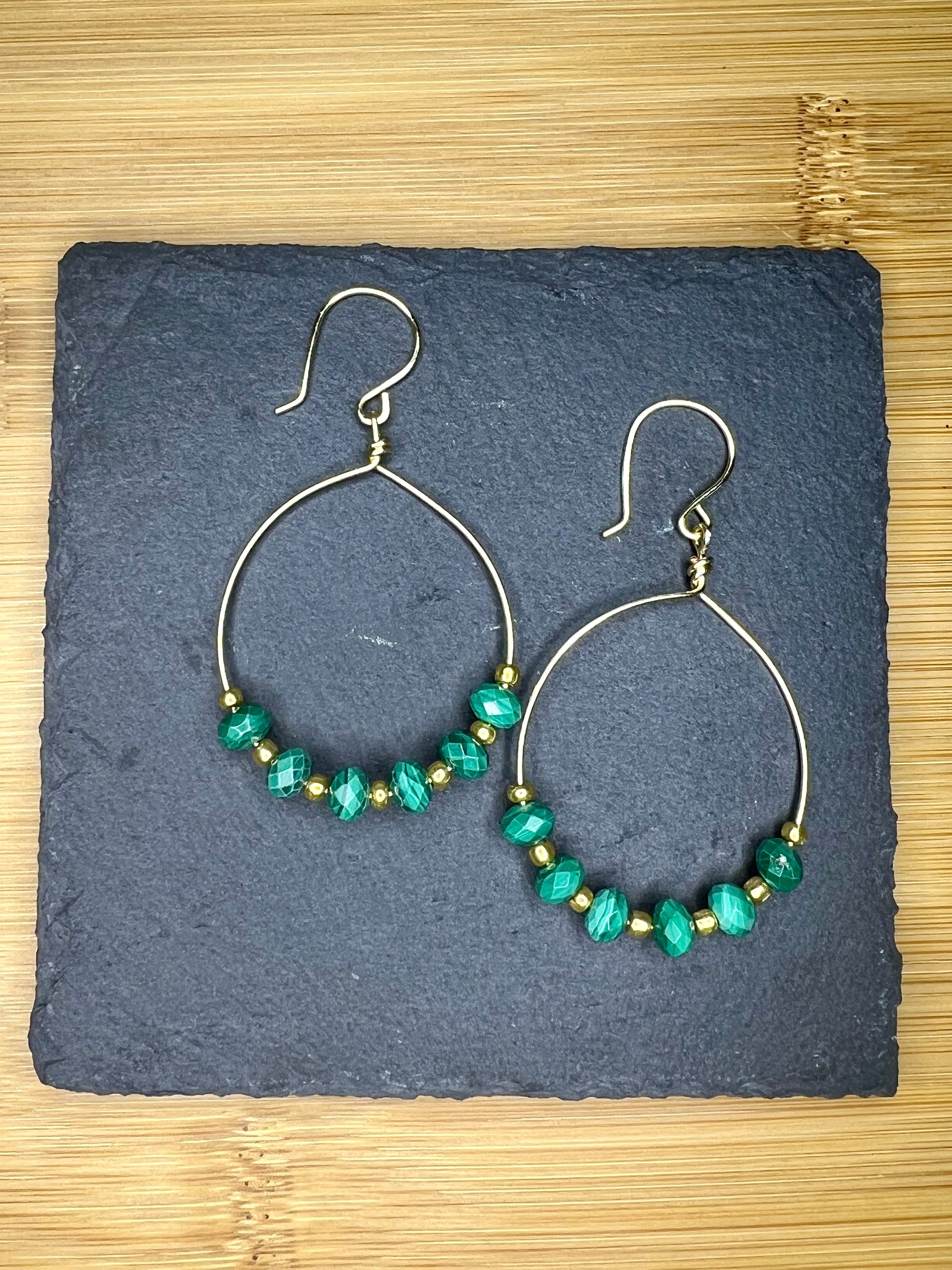 A pair of gold hoop earrings with green beads, displayed on a dark slate surface.