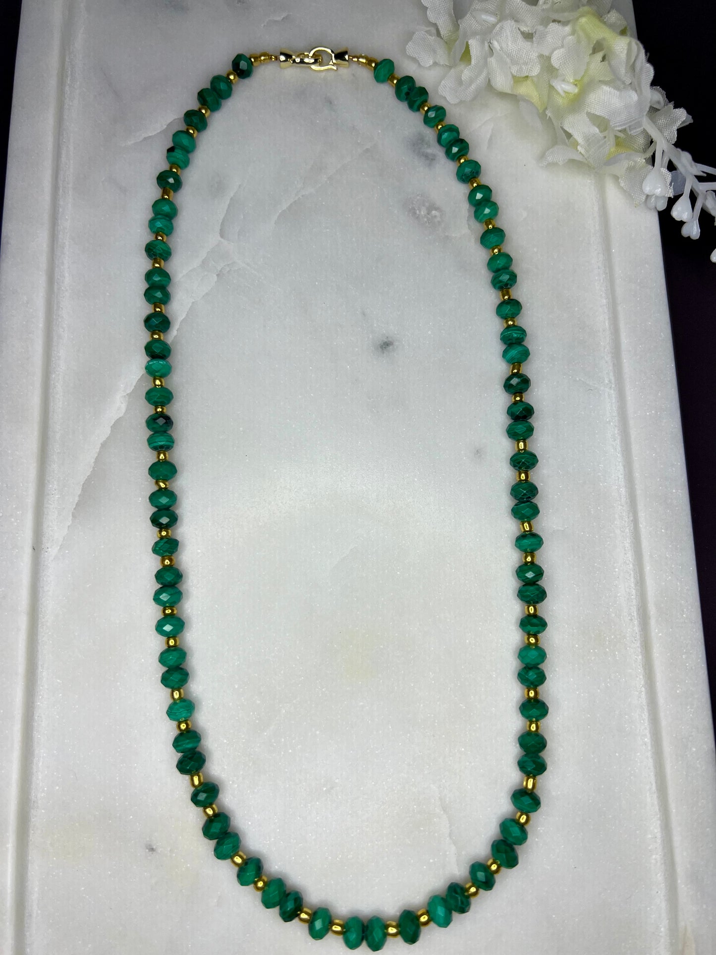 A necklace with green beads and gold beads is displayed on a white marble surface, accompanied by a small white flower.