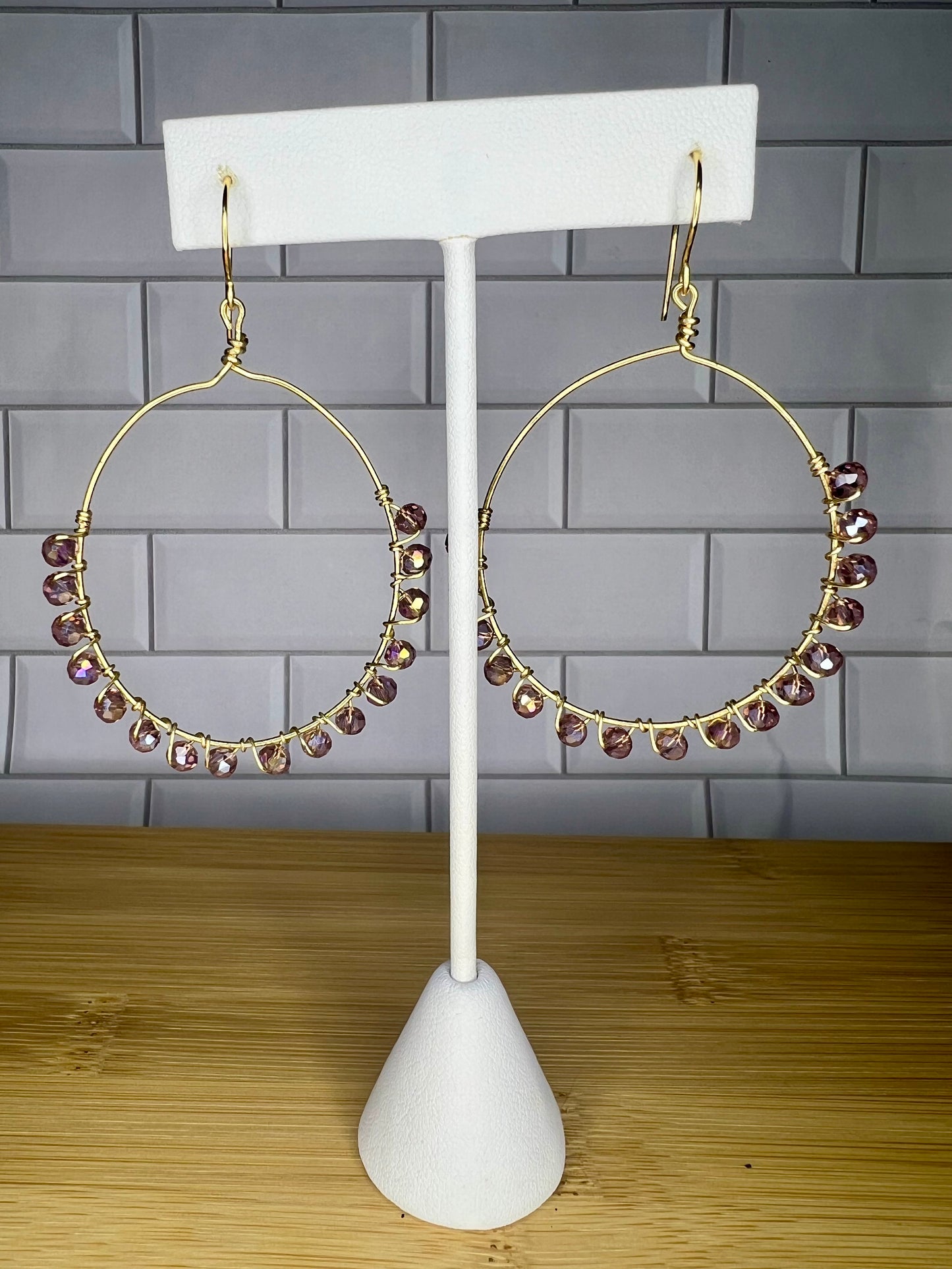 A pair of gold hoop earrings with small purple gemstones hanging from them, displayed on a white earring stand against a gray tiled wall.