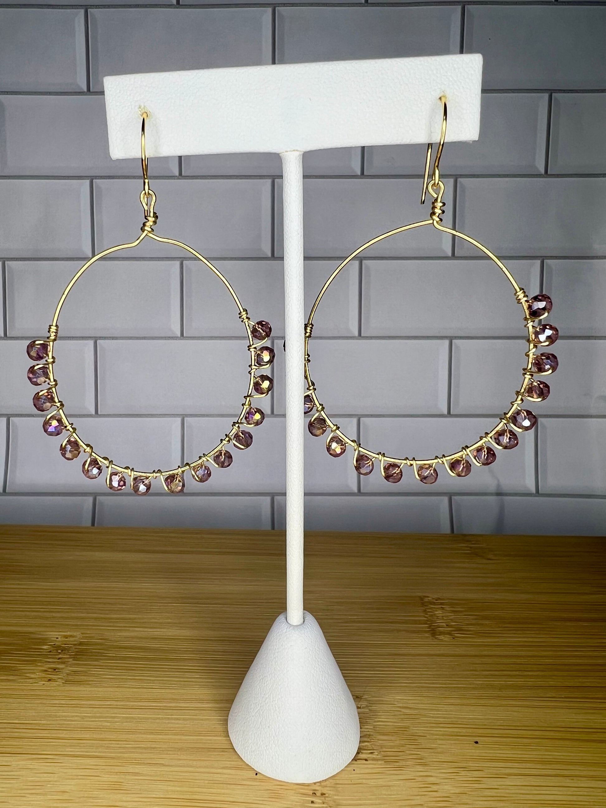 A pair of gold hoop earrings with small purple gemstones hanging from them, displayed on a white earring stand against a gray tiled wall.