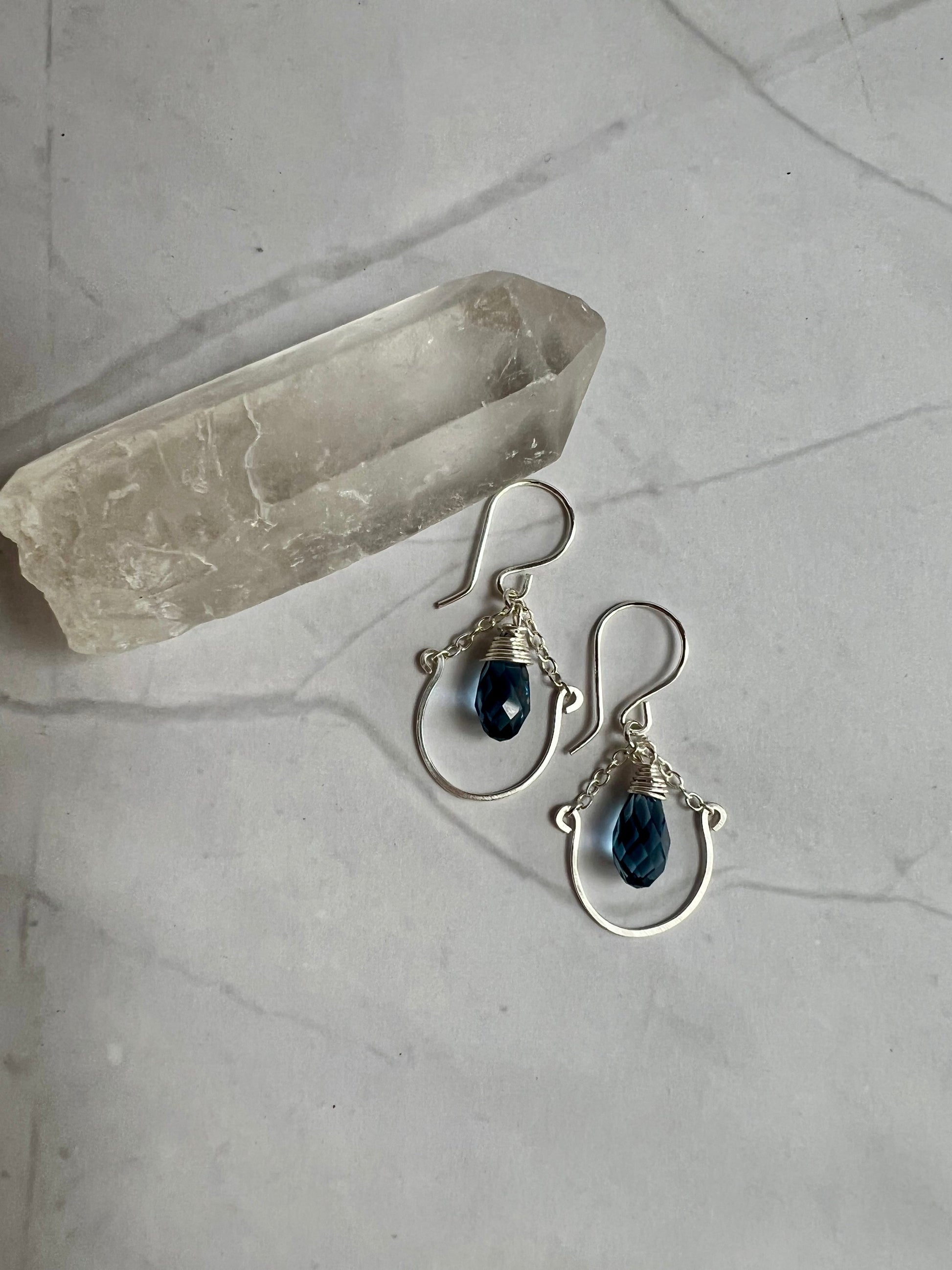 A pair of silver earrings with blue gemstones, resting on a white marble surface next to a crystal.