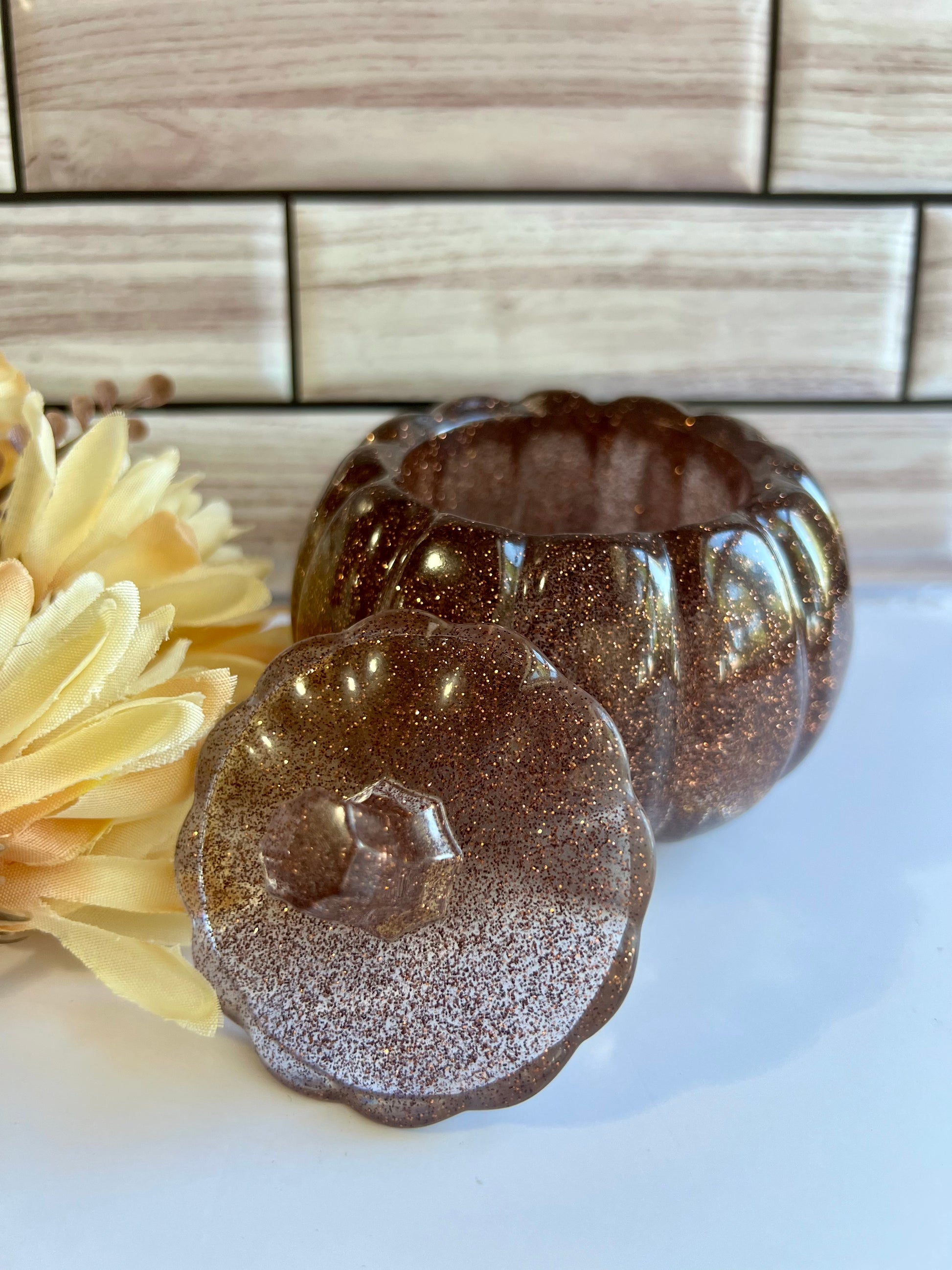 a brown glass bowl with a lid, placed on a white surface next to a yellow flower.