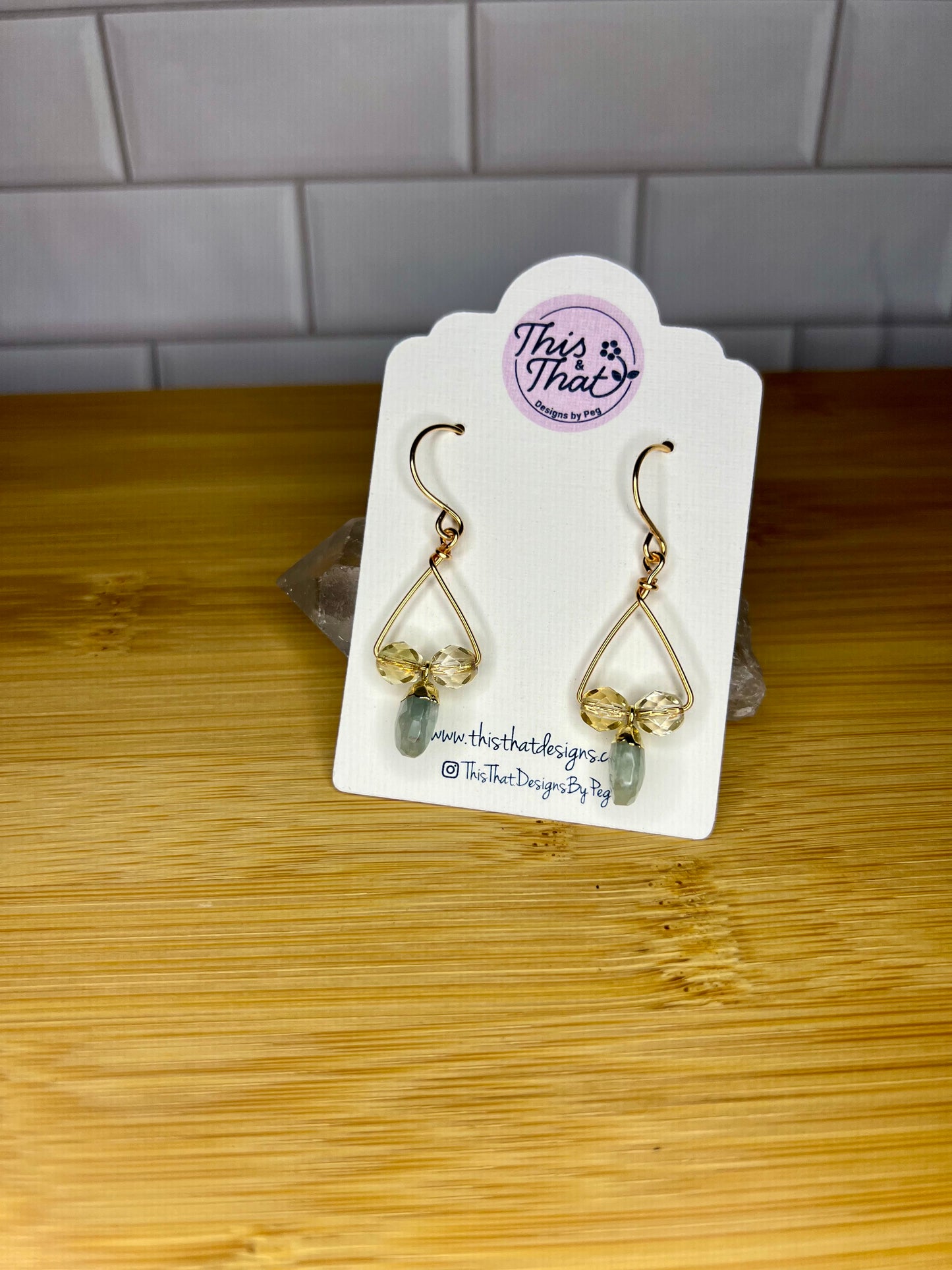 A pair of gold earrings with green stones hanging from them, displayed on a white card with a logo and text.
