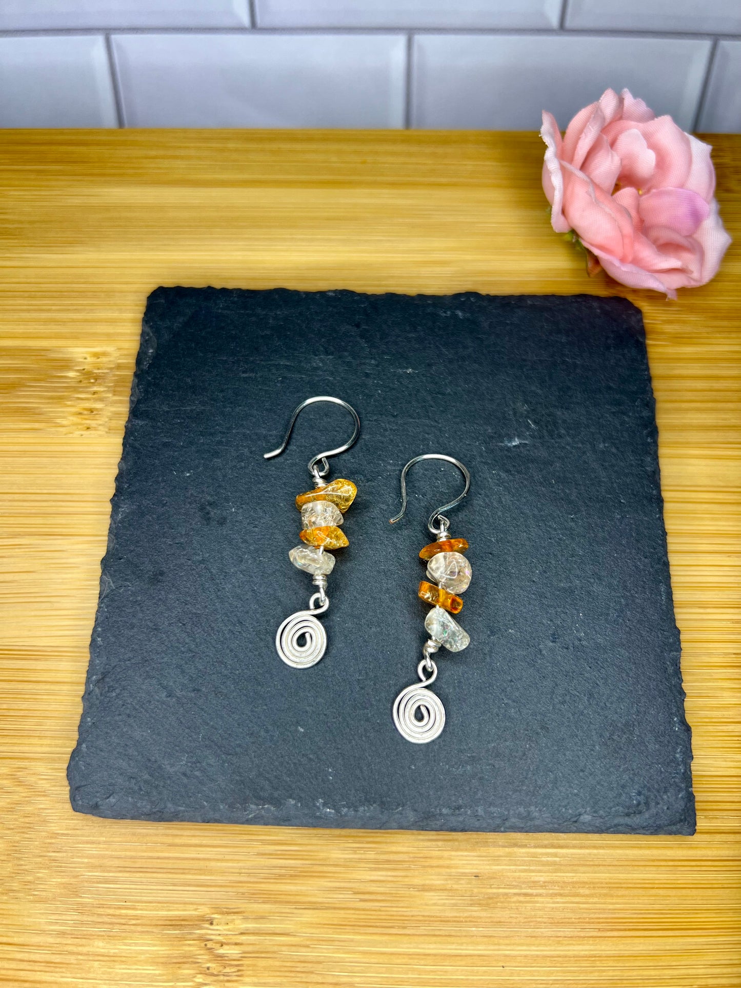 A pair of silver earrings with colorful stones dangles from the hooks, placed on a black slate or slate board. A single pink flower is also present in the image.