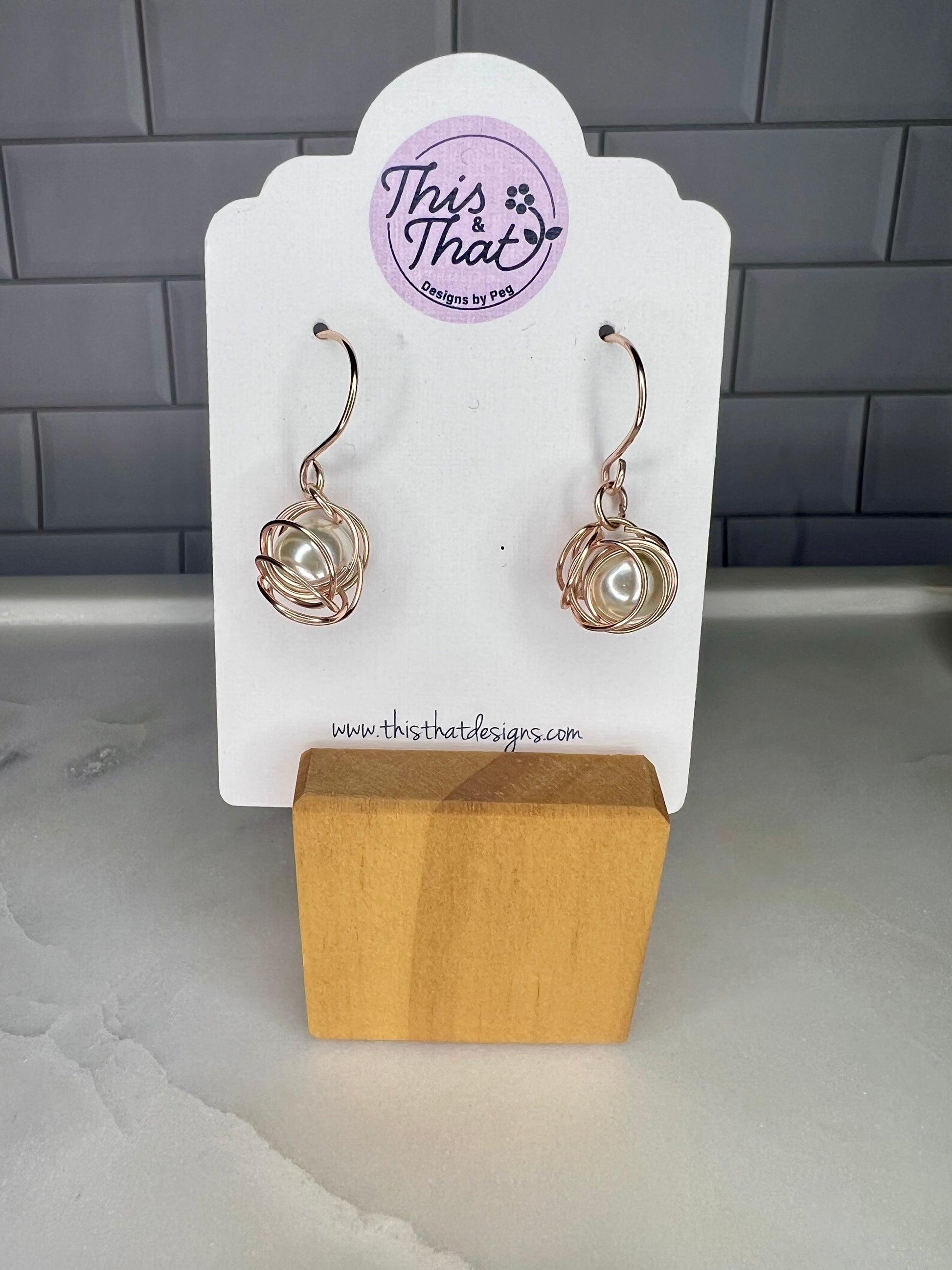 This image shows a pair of earrings displayed on a white card stand. The earrings are made of metal and have a unique, twisted design. The card stand is made of wood and has a website address printed on it.